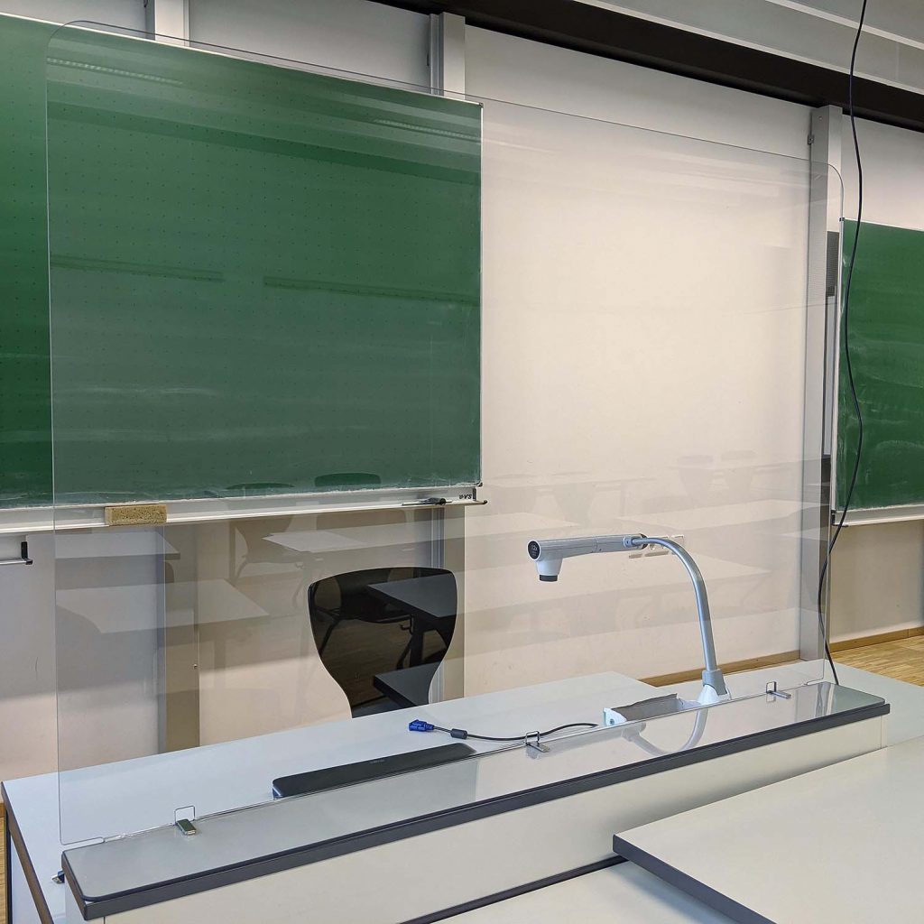 Infektionsschutzwand aus unzerbrechlichem Polycarbonat in einem Klassenzimmer, fest montiert auf dem Lehrerpult. Entwickelt und produziert von RIWA Display GmbH in der Universitätsstadt Garching bei München.